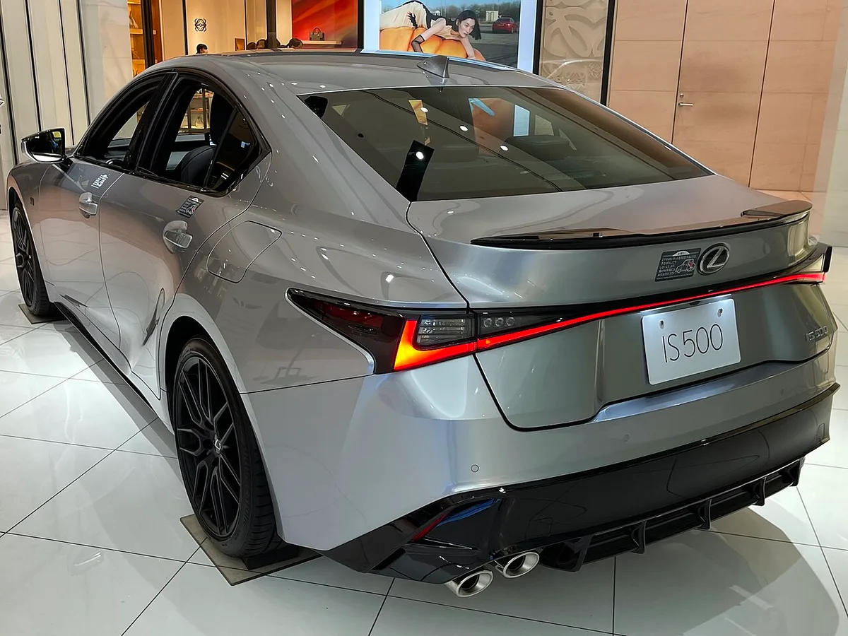 Rear three-quarter view of a silver Lexus IS 500 F Sport Performance in a showroom, showing its stacked exhaust tips and black rear lip spoiler