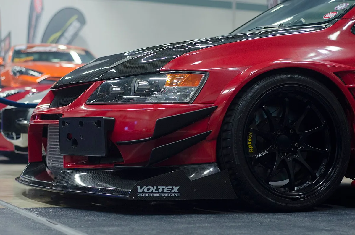 Front corner of a red Mitsubishi Lancer Evolution IX fitted with a Voltex aero kit, showing its carbon front splitter, canards, and vented fender